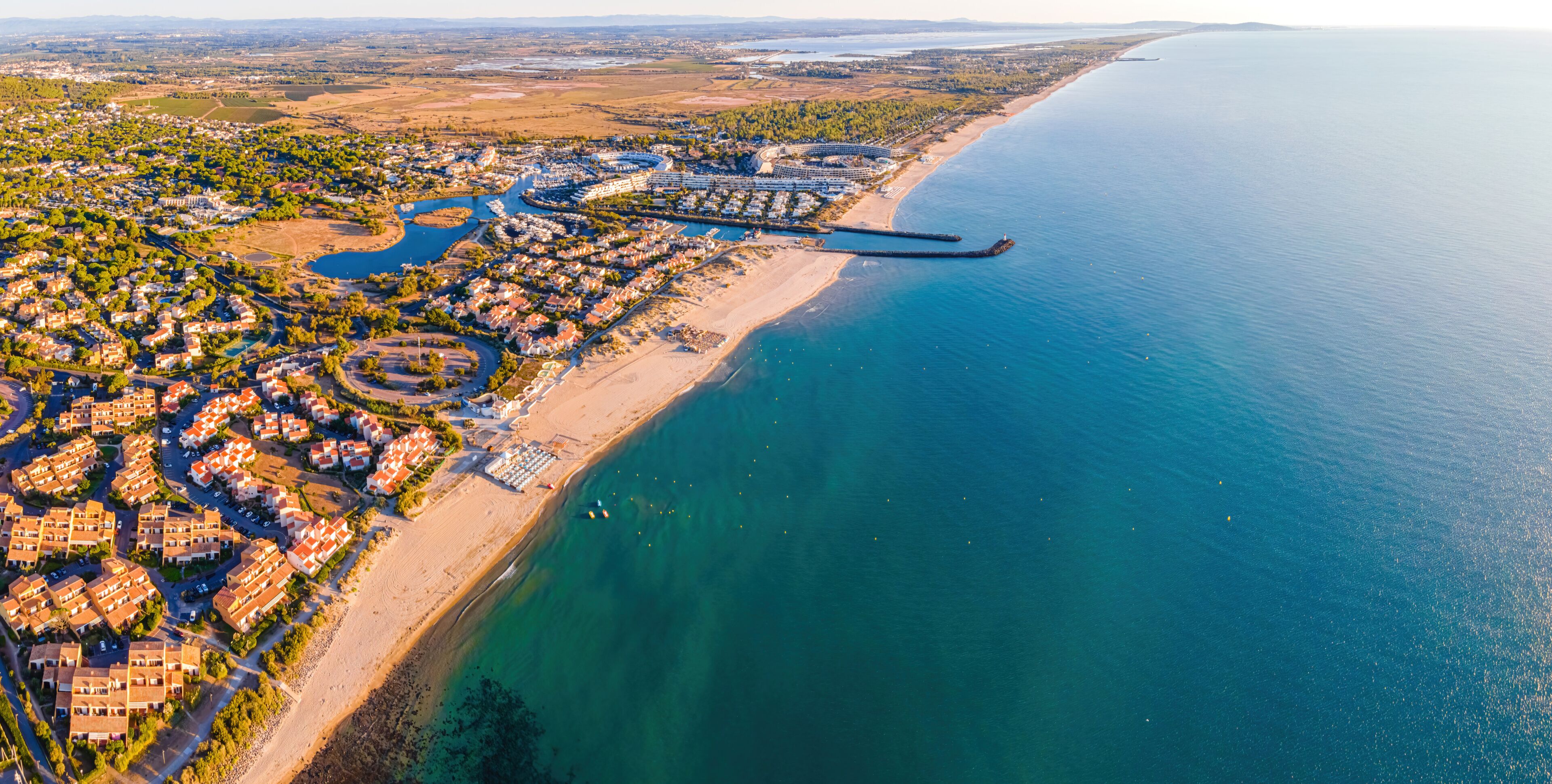 An aerial view of a coastal town with a marina, residential areas, and a long beach.