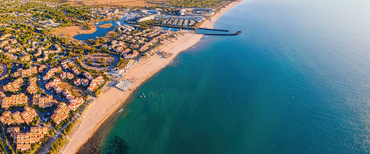 An aerial view of a coastal town with a marina, residential areas, and a long beach.