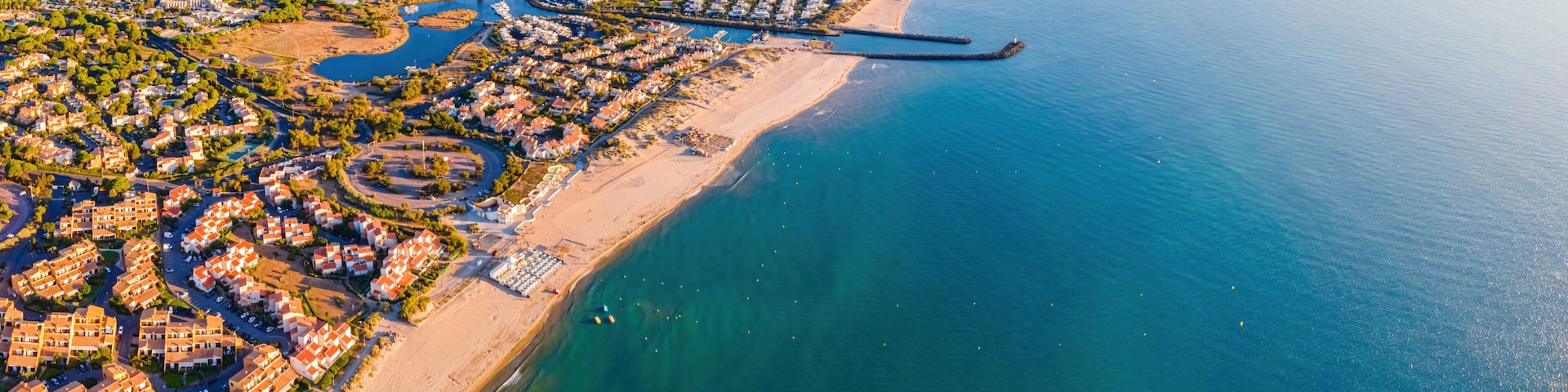 An aerial view of a coastal town with a marina, residential areas, and a long beach.