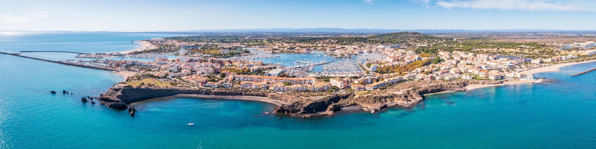 An aerial view of a coastal town with a marina, rocky coastline, and surrounding hills.