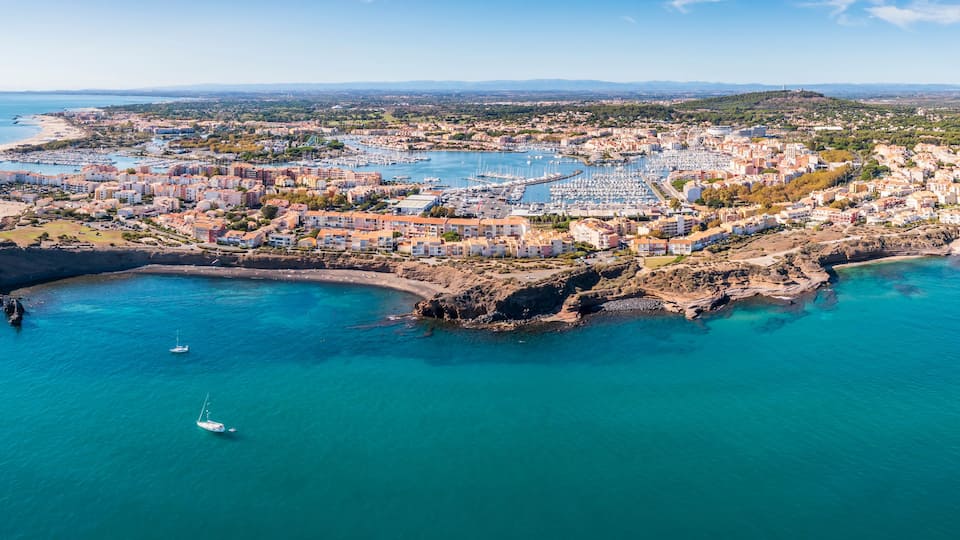 An aerial view of a coastal town with a marina, rocky coastline, and surrounding hills.