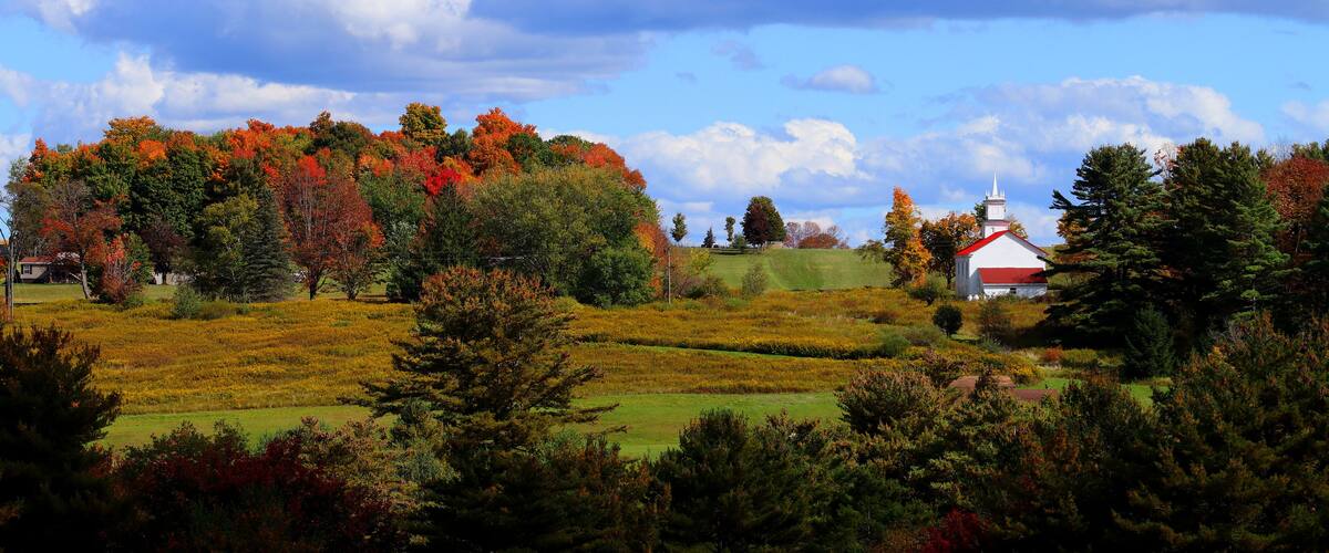 A rural landscape with a white church, autumn-colored trees, and a clear blue sky.