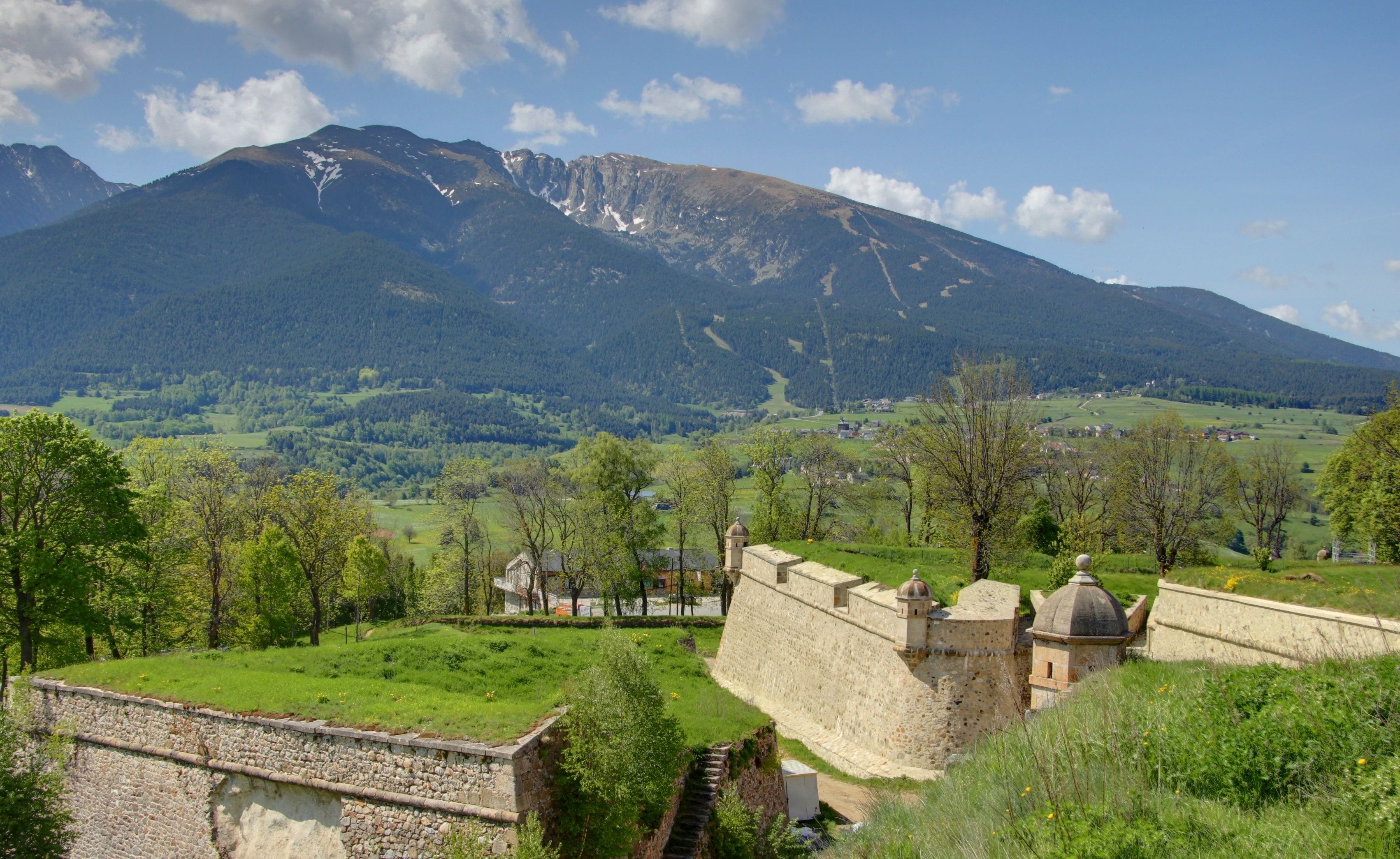 A castle with a stone wall and a tower, surrounded by green fields and trees, with a mountain in the background.