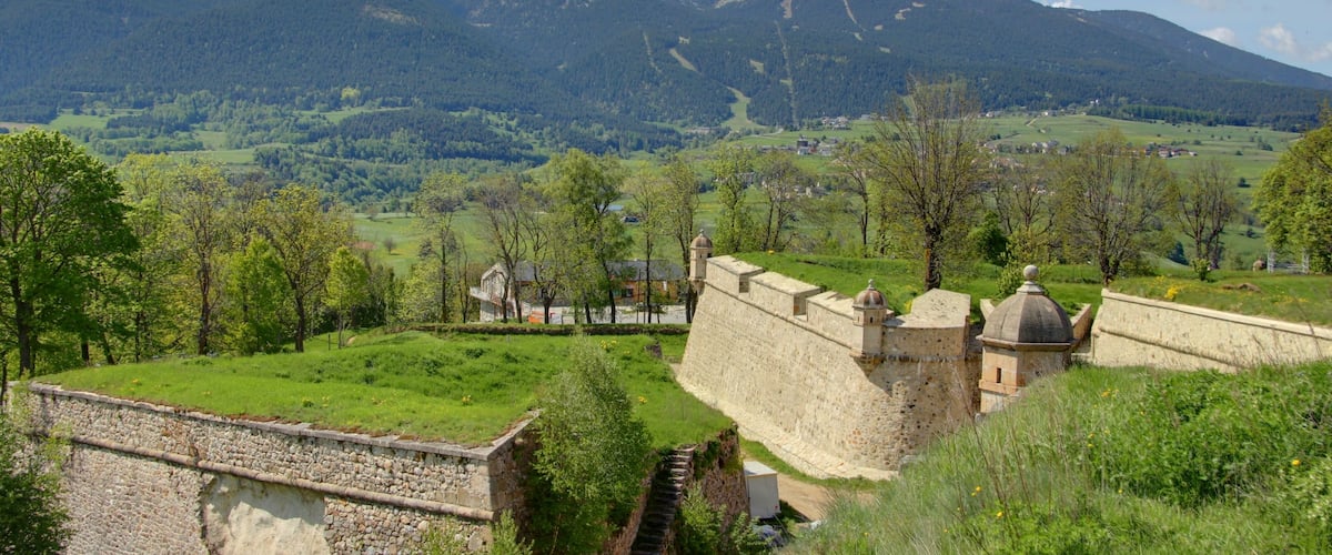 Un castillo con muro de piedra y una torre, rodeado de campos verdes y árboles, con una montaña al fondo.