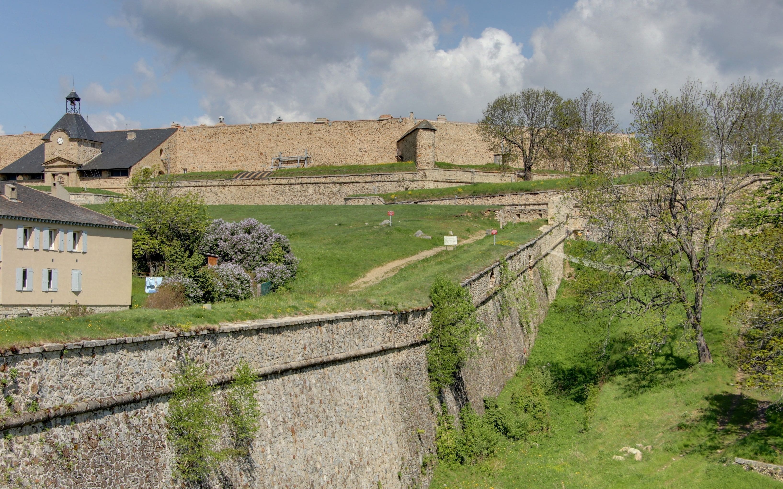 A stone fortification with a watchtower, a house with shutters, and a tree with purple flowers.