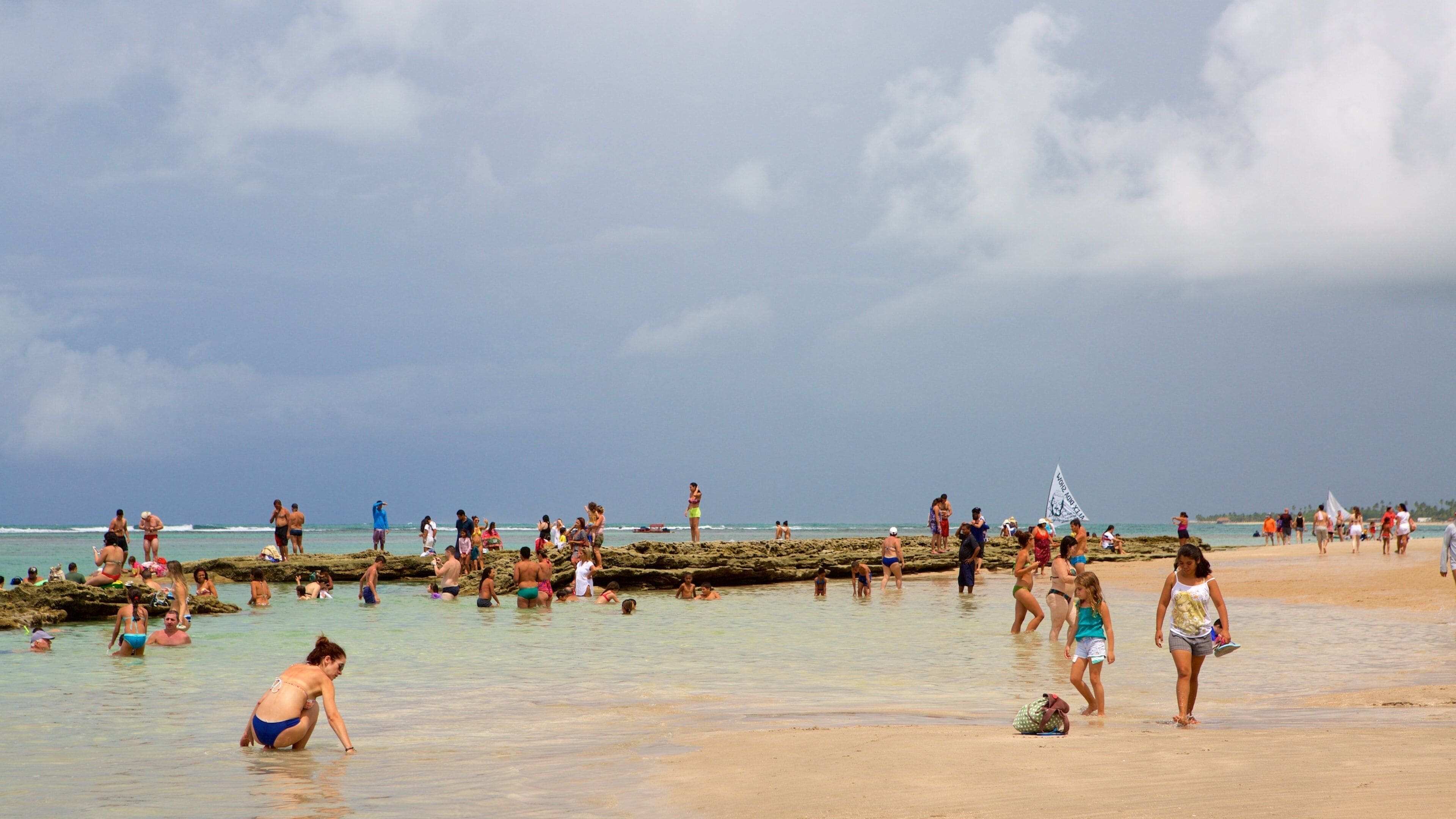 Una playa concurrida con gente en el agua y en la arena, bajo un cielo nublado.