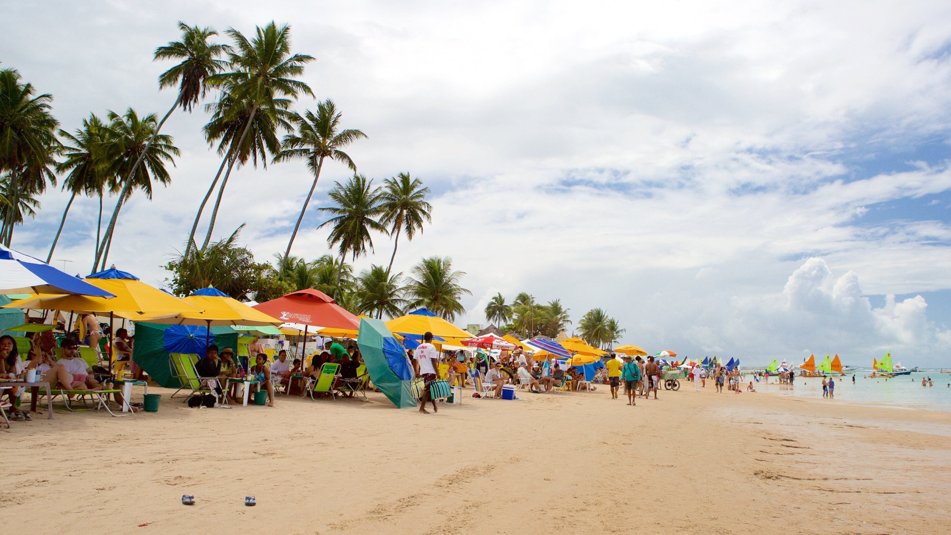 Ein voller Strand mit Palmen und bunten Sonnenschirmen.