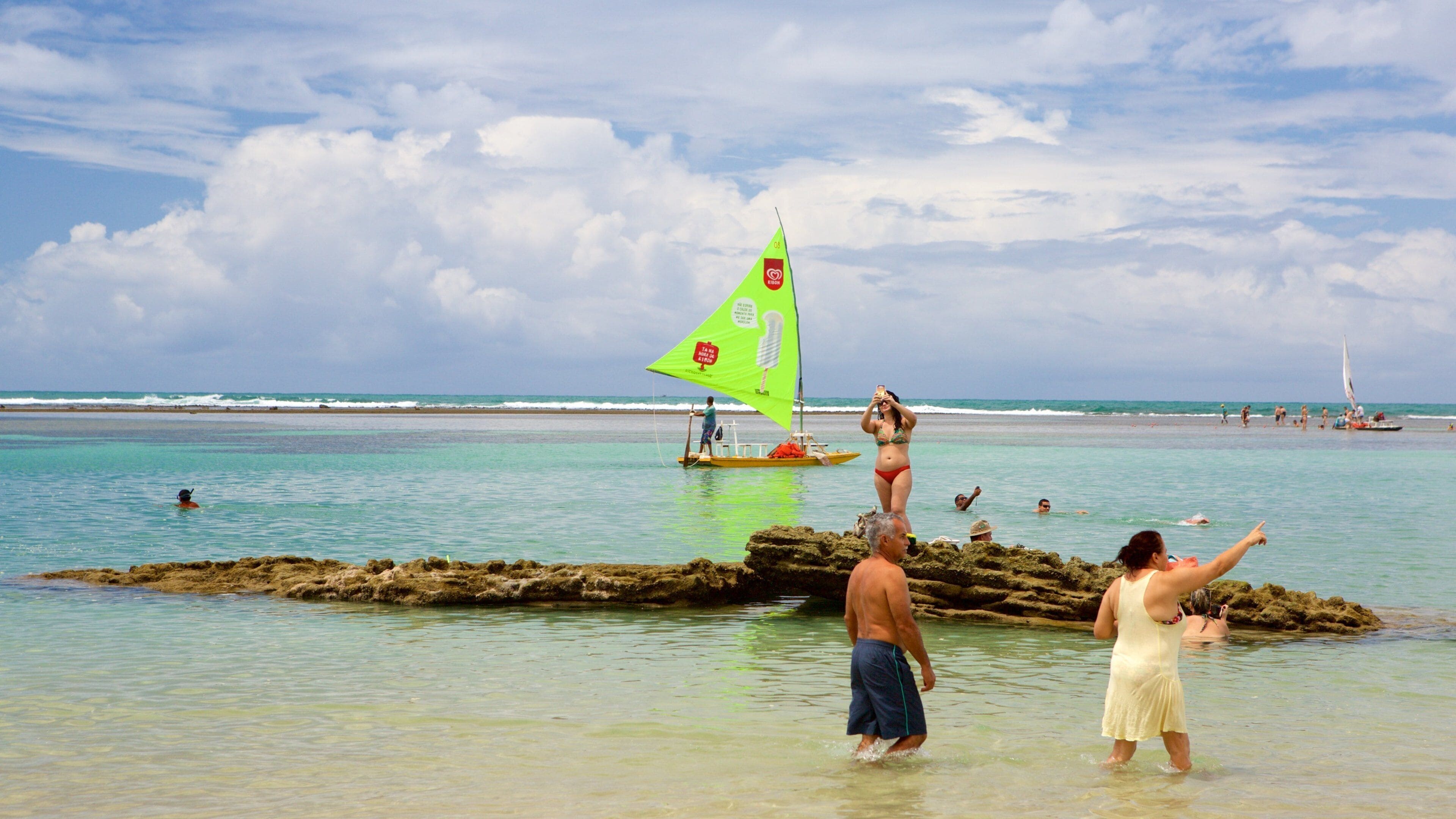 Una playa con agua turquesa cristalina, un velero y gente nadando y caminando en el agua.