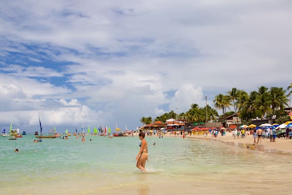 Praia lotada com pessoas nadando e tomando sol, coqueiros e guarda-sóis coloridos.