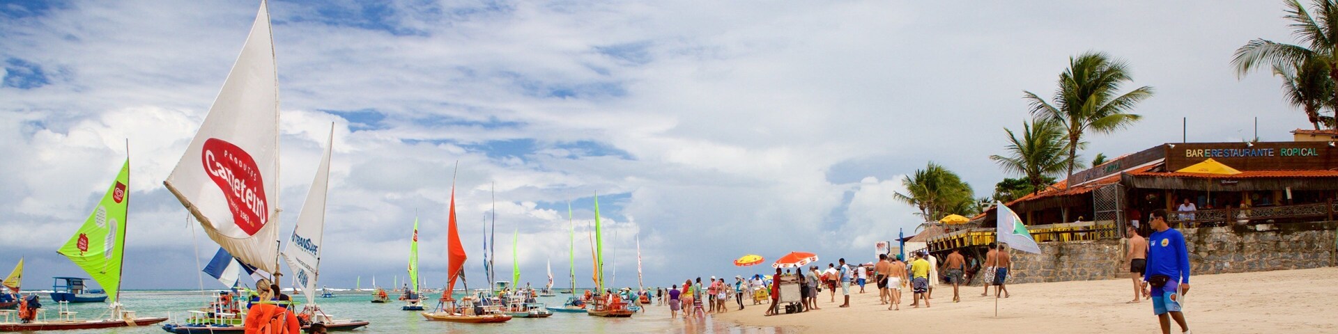 Uma praia com veleiros, pessoas e um restaurante.