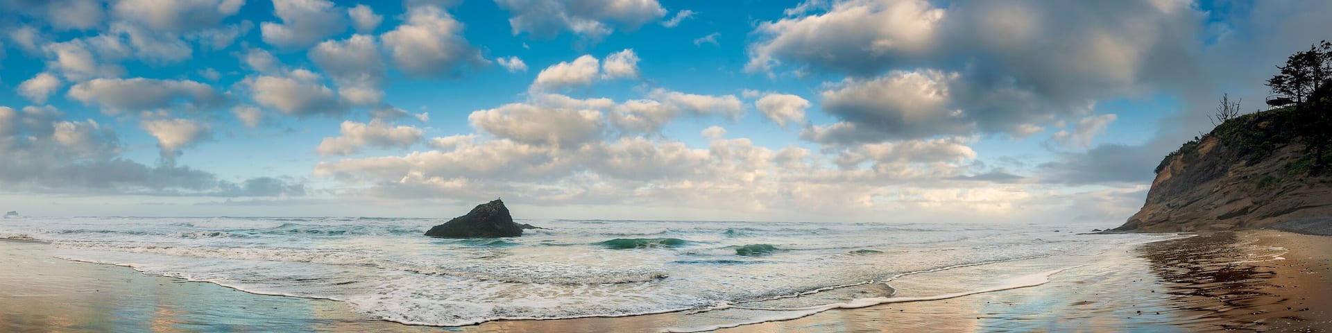 A beach with waves, a rocky outcrop, and a cloudy sky.