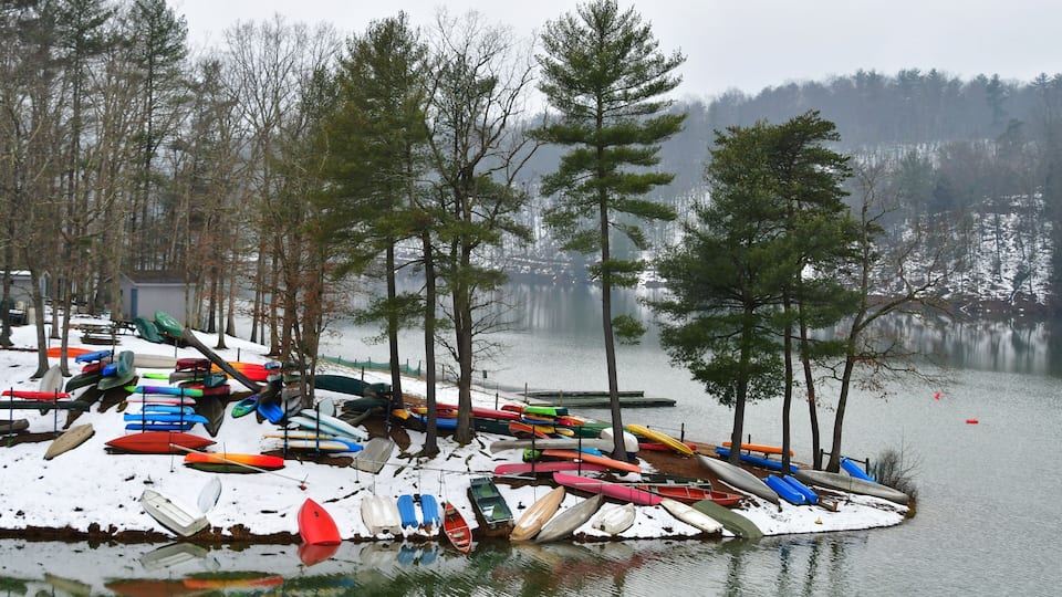 A snowy lakeside with numerous colorful kayaks and canoes docked along the shore.