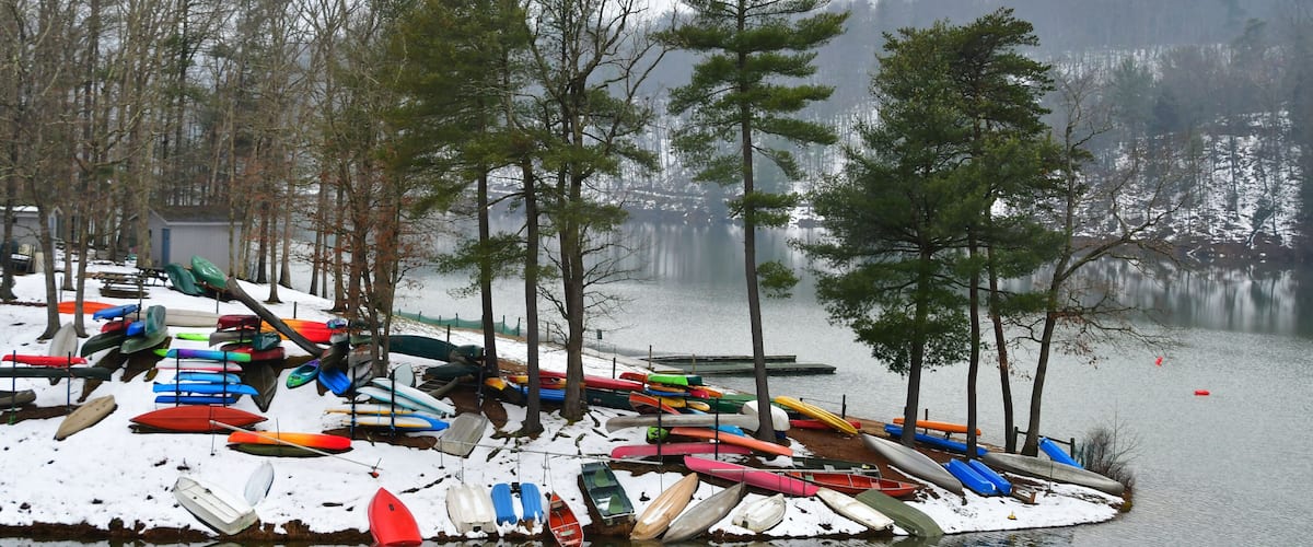 A snowy lakeside with numerous colorful kayaks and canoes docked along the shore.