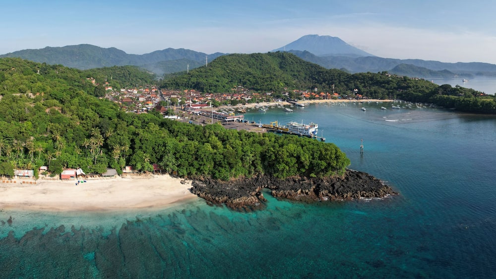 A coastal town with a beach, clear turquoise waters, and lush green hills in the background.