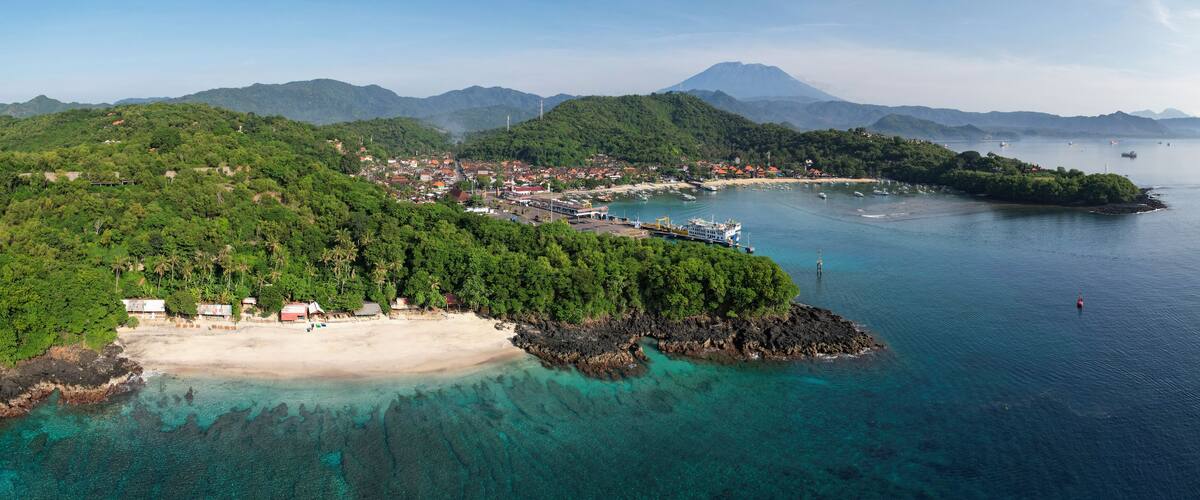 A coastal town with a beach, clear turquoise waters, and lush green hills in the background.