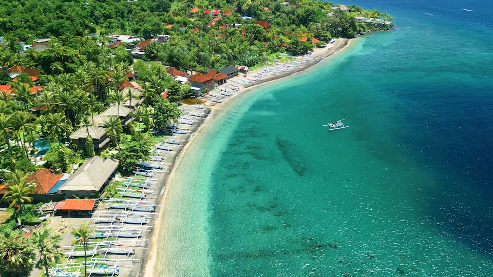 A coastal area with a clear turquoise sea, a row of beachside houses, and a line of boats along the shore.