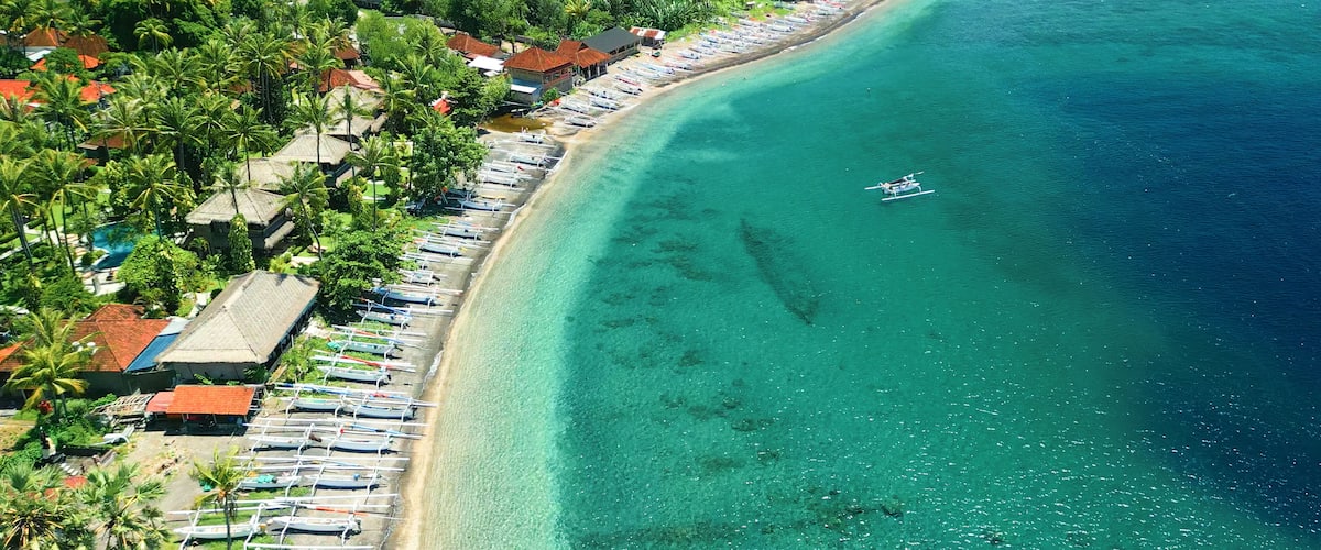 A coastal area with a clear turquoise sea, a row of beachside houses, and a line of boats along the shore.