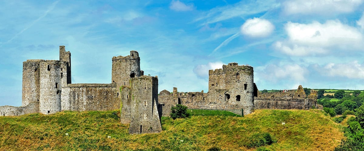 A historic stone castle with multiple towers and walls, situated on a grassy hill under a partly cloudy sky.