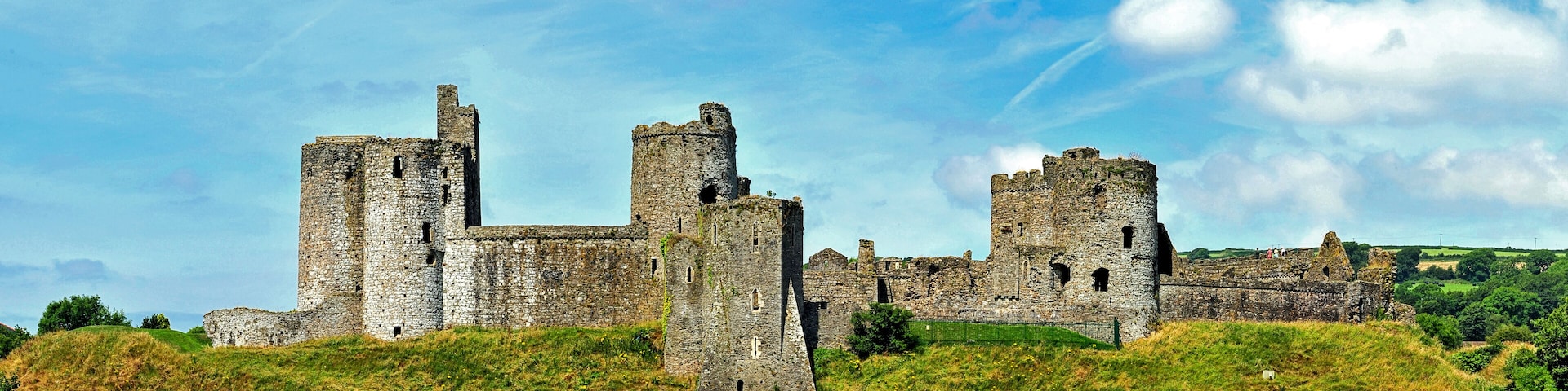 A historic stone castle with multiple towers and walls, situated on a grassy hill under a partly cloudy sky.