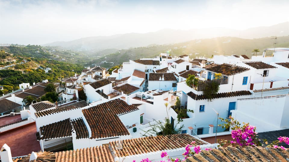 A coastal village with white buildings and terracotta roofs, surrounded by greenery and mountains.