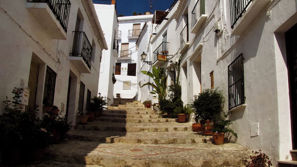 A narrow cobblestone street flanked by white buildings with black railings, leading up a set of stone steps.