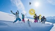 A group of people snowboarding on a snowy mountain with a clear blue sky and snow-covered peaks in the background.