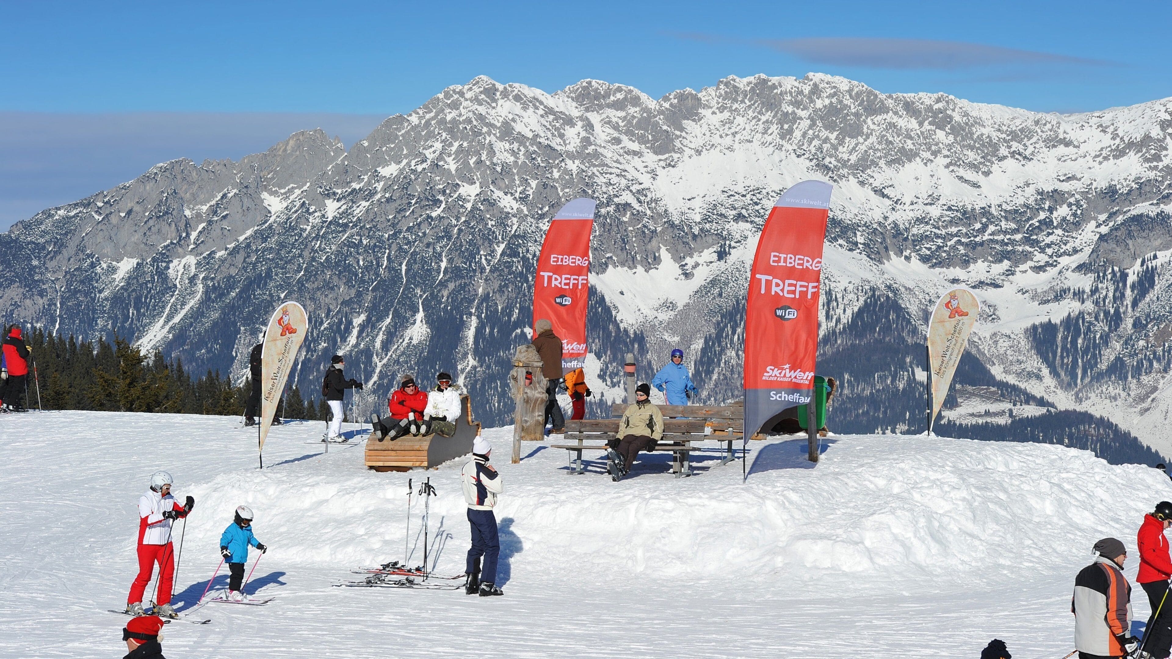 Skiers on a snowy mountain with red banners and a wooden bench.