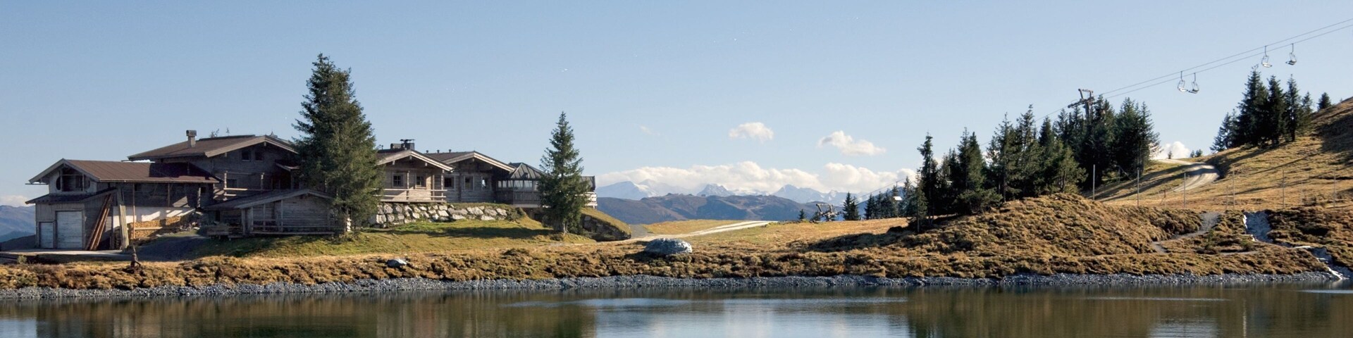 Blick auf einen See mit einem Holzhäuschen, einer Seilbahn und schneebedeckten Bergen im Hintergrund.