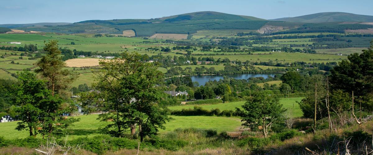 A lush green valley with rolling hills, a lake, and scattered houses.