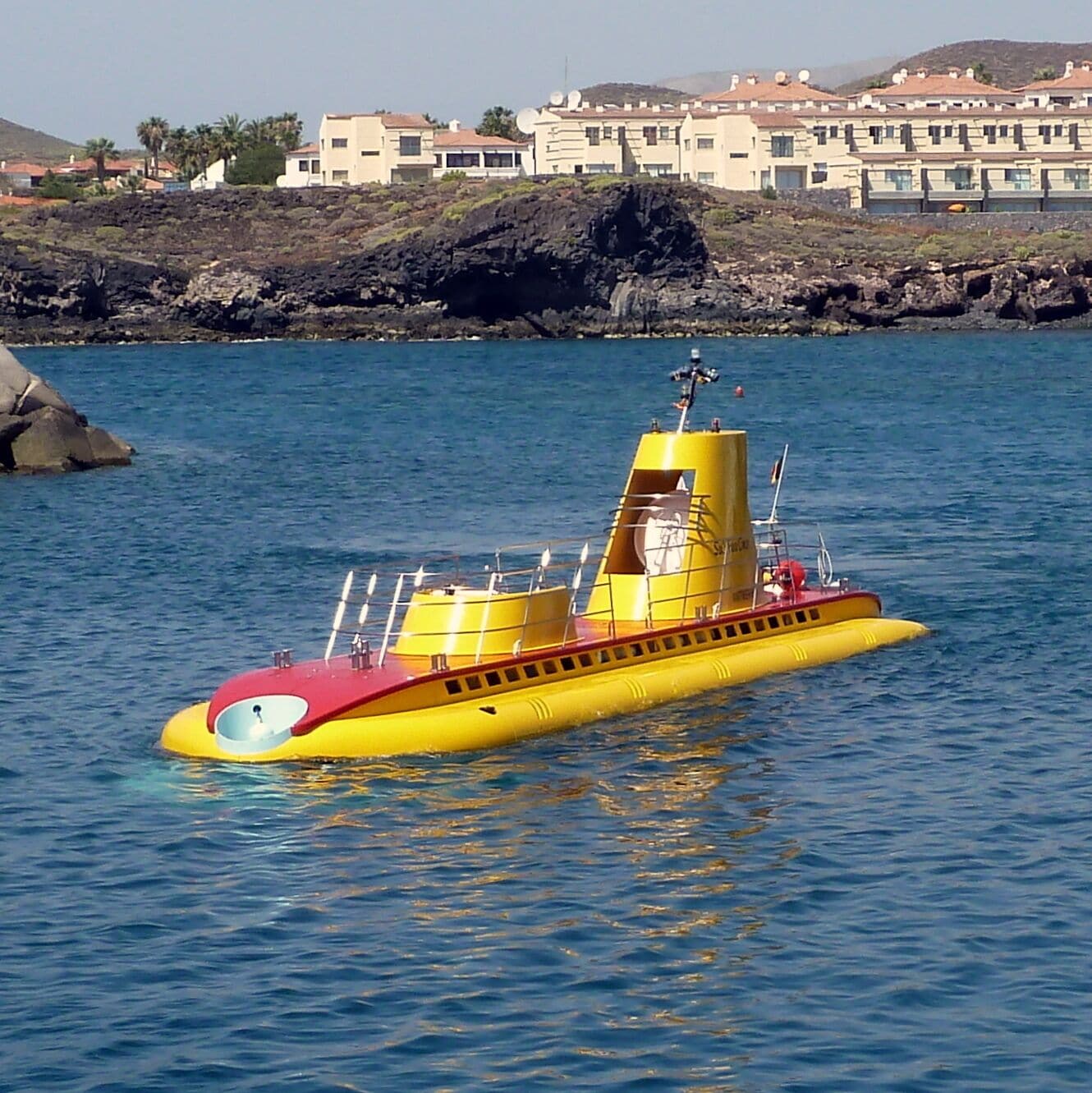 A yellow submarine in calm waters with a rocky shoreline and buildings in the background.