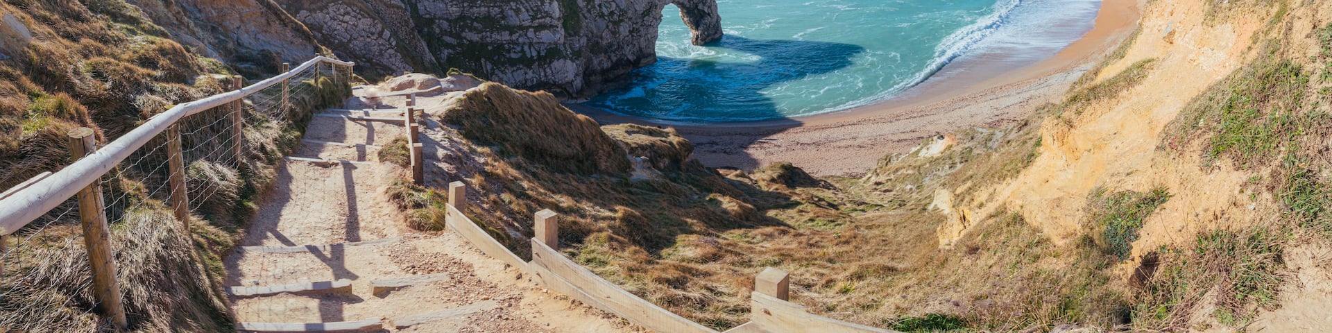 A coastal landscape with a prominent rock arch formation, a sandy beach, and a clear blue sky.