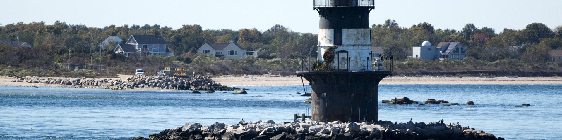 Un faro en una pequeña isla con costa rocosa, rodeado de casas y árboles.