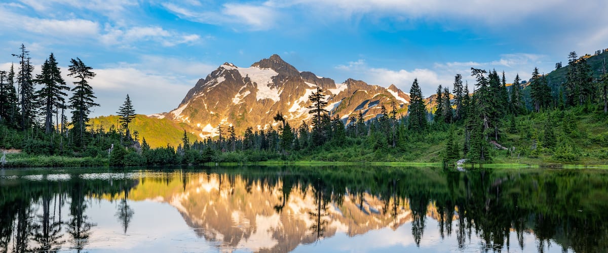 A mountain range with snow-capped peaks, a calm lake reflecting the scenery, and a forested area with tall trees.