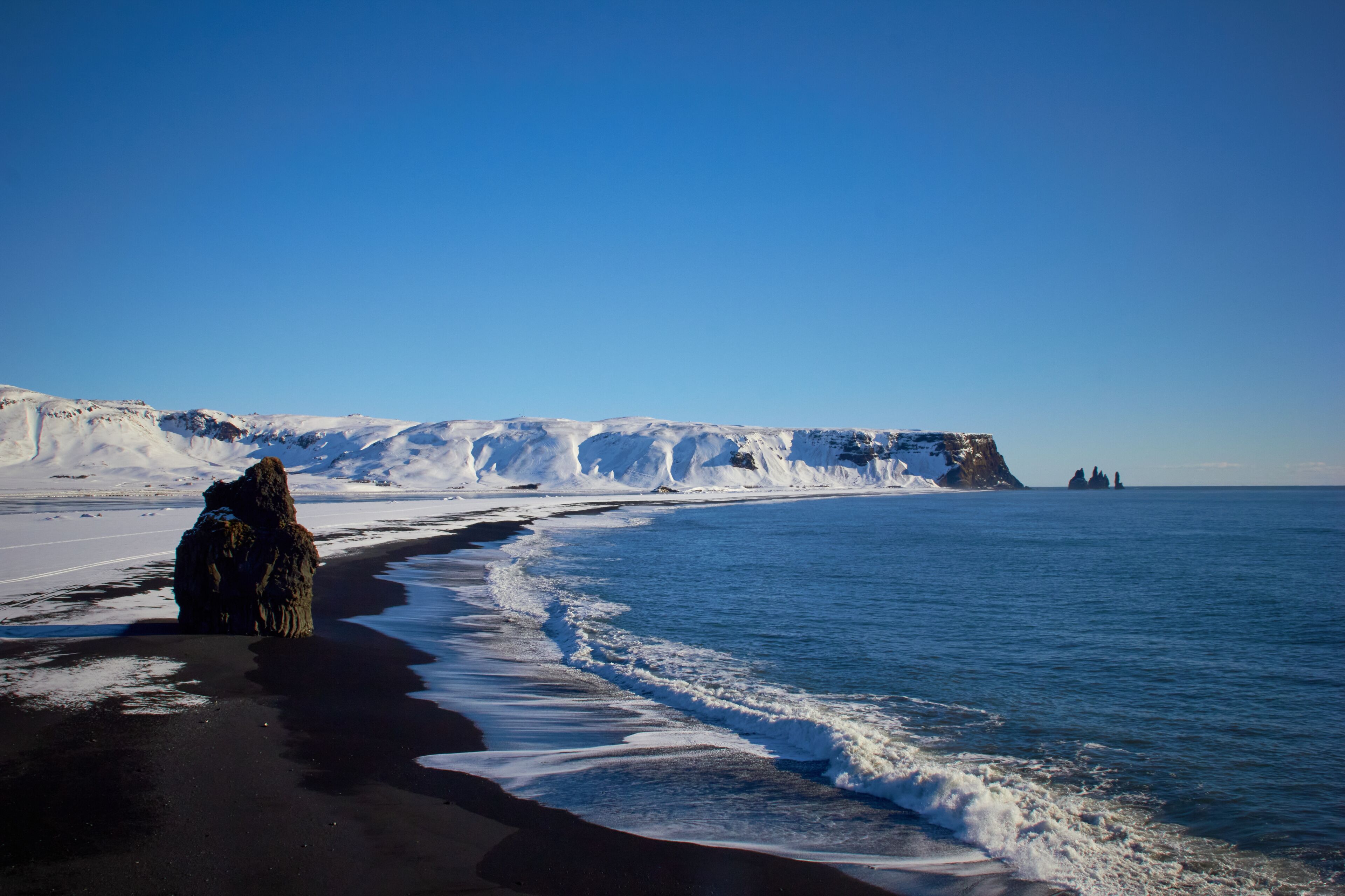 A snowy beach with a large rock formation and cliffs in the background.