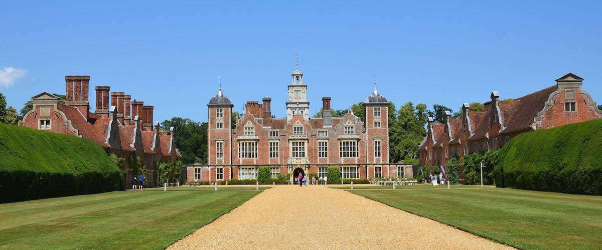 A large, historic brick building with a central tower, surrounded by well-manicured lawns and a gravel path leading to the entrance.
