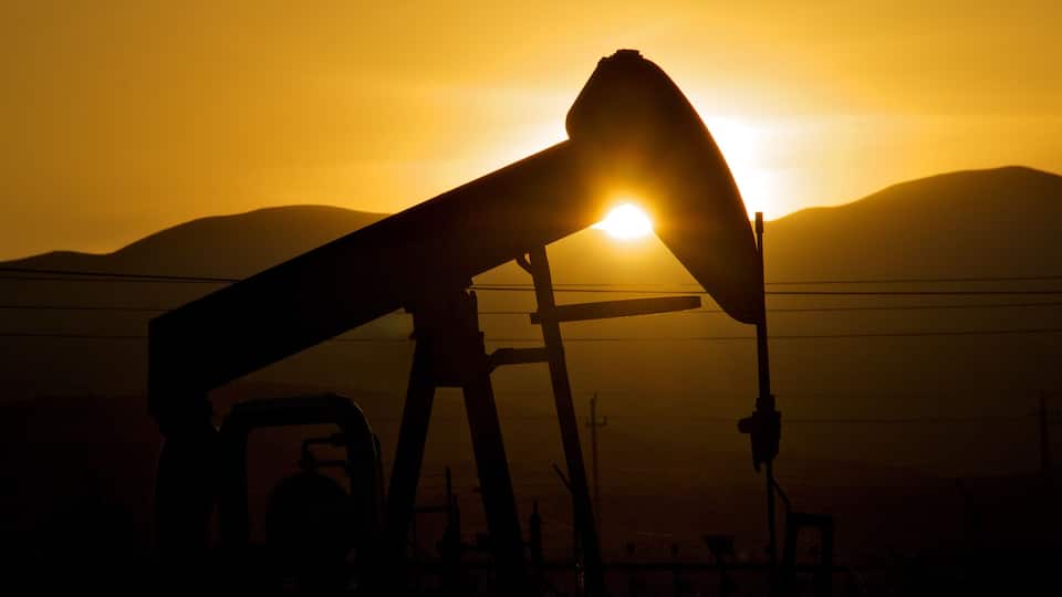 A silhouette of an oil pumpjack against a sunset with mountains in the background.