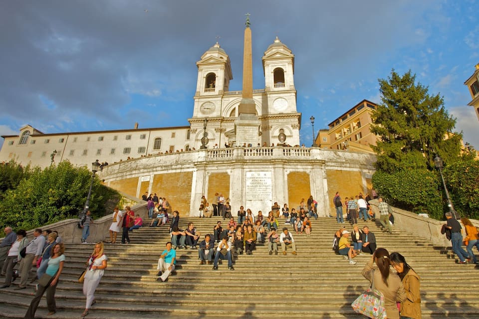 A historic church with twin bell towers and a central obelisk, surrounded by a crowd of people on the steps.
