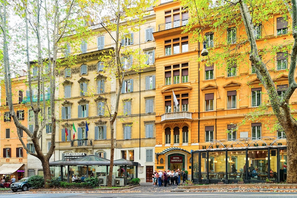 Ein historisches Gebäude mit gelber Fassade, ein Café und eine Bushaltestelle mit Wartenden.