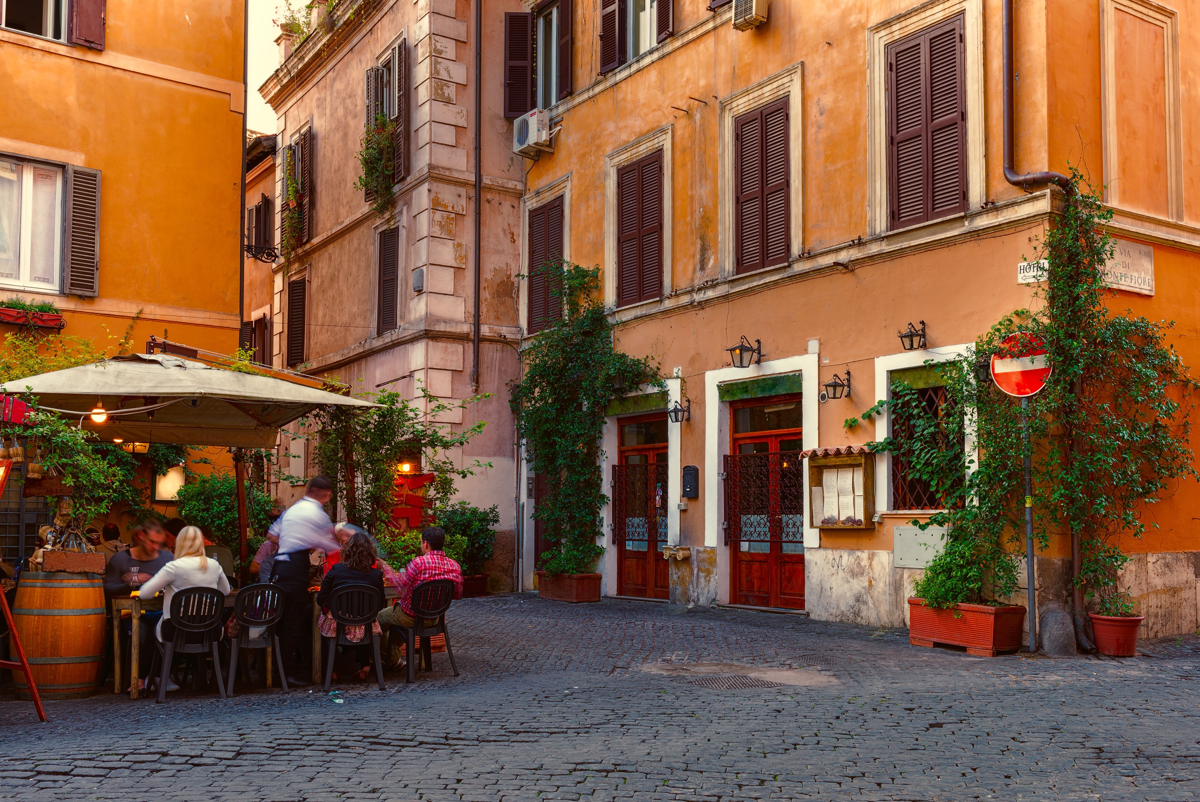 A cobblestone street with outdoor seating at a restaurant, surrounded by historic buildings with shutters and potted plants.