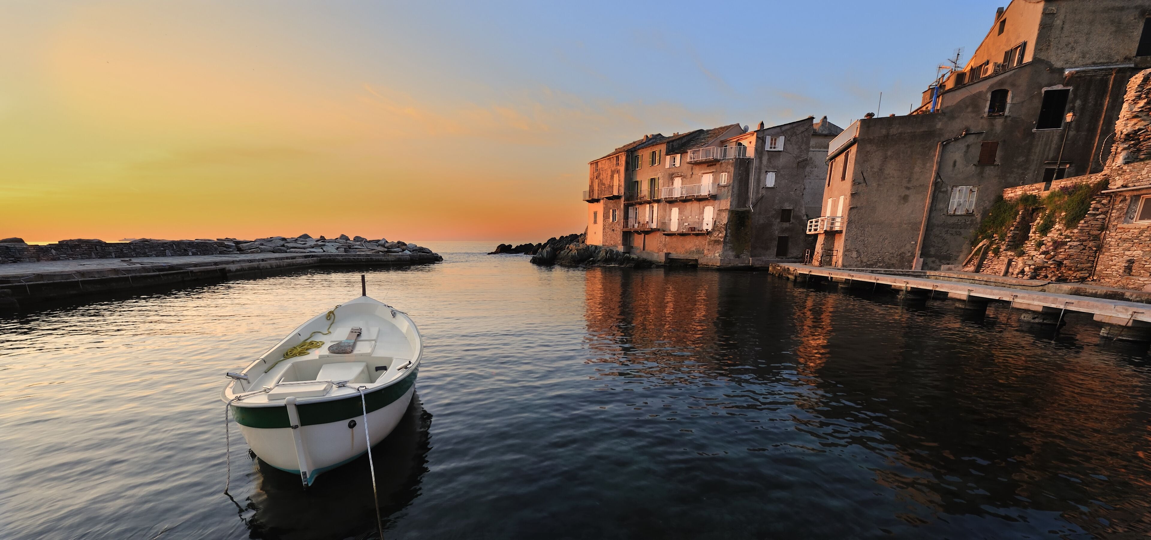 A calm harbor with a small boat docked near a stone pier, surrounded by old buildings with balconies.
