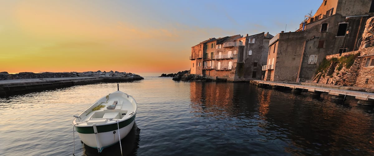 A calm harbor with a small boat docked near a stone pier, surrounded by old buildings with balconies.