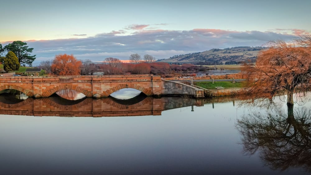 A stone bridge over a calm river, surrounded by autumn trees and a distant church.
