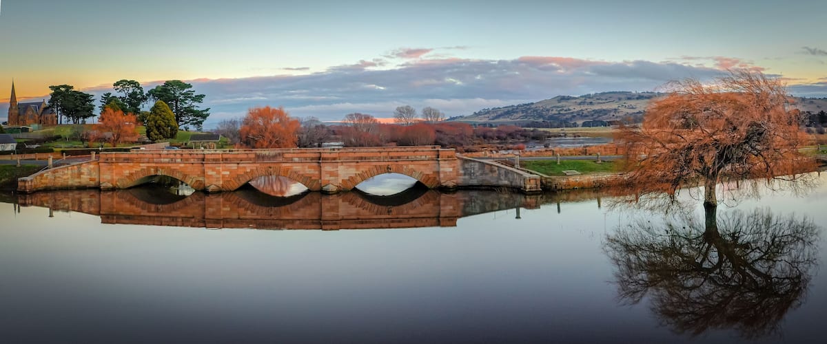 A stone bridge over a calm river, surrounded by autumn trees and a distant church.