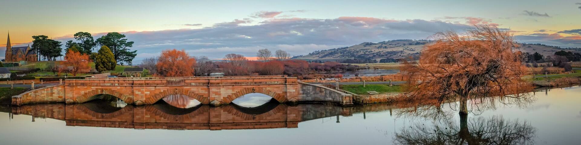 A stone bridge over a calm river, surrounded by autumn trees and a distant church.
