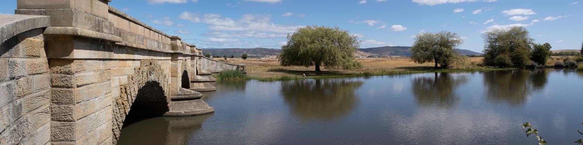 Uma ponte de pedra sobre um rio calmo, rodeada por árvores e colinas ao longe.