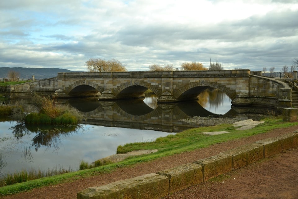A stone bridge over a calm river with grassy banks and a cloudy sky.