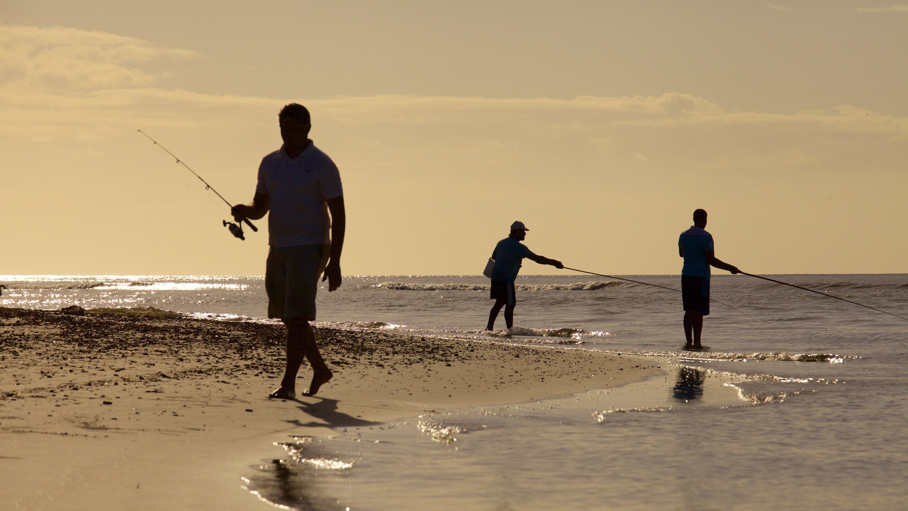 Three people fishing on a beach during sunset.