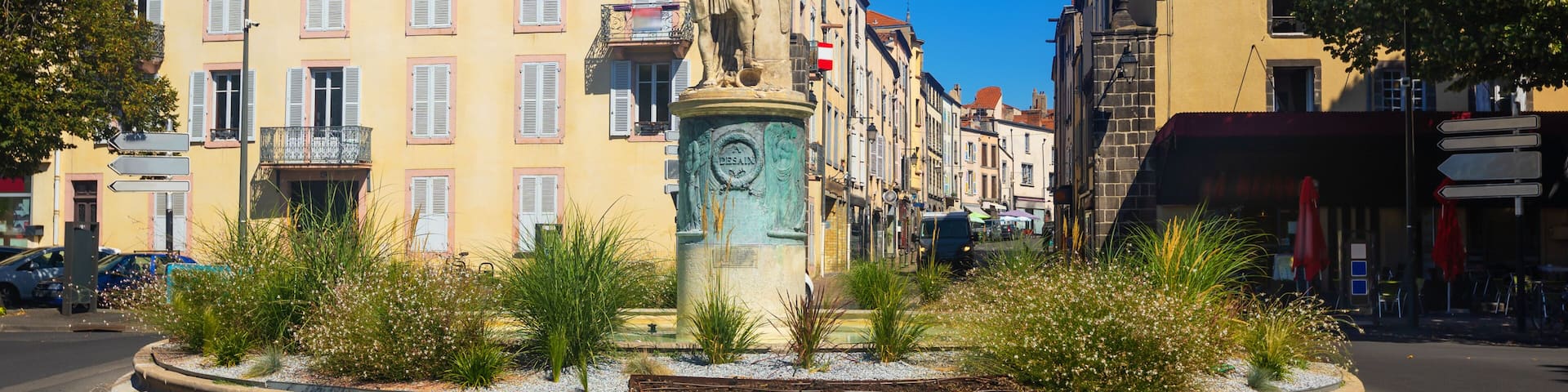 A roundabout with a statue, surrounded by buildings with pastel-colored facades and red-tiled roofs.