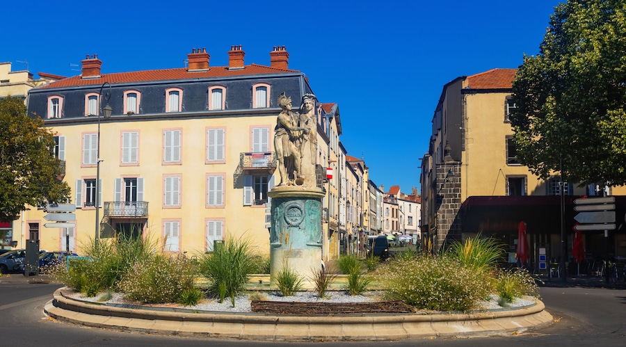 A roundabout with a statue, surrounded by buildings with pastel-colored facades and red-tiled roofs.
