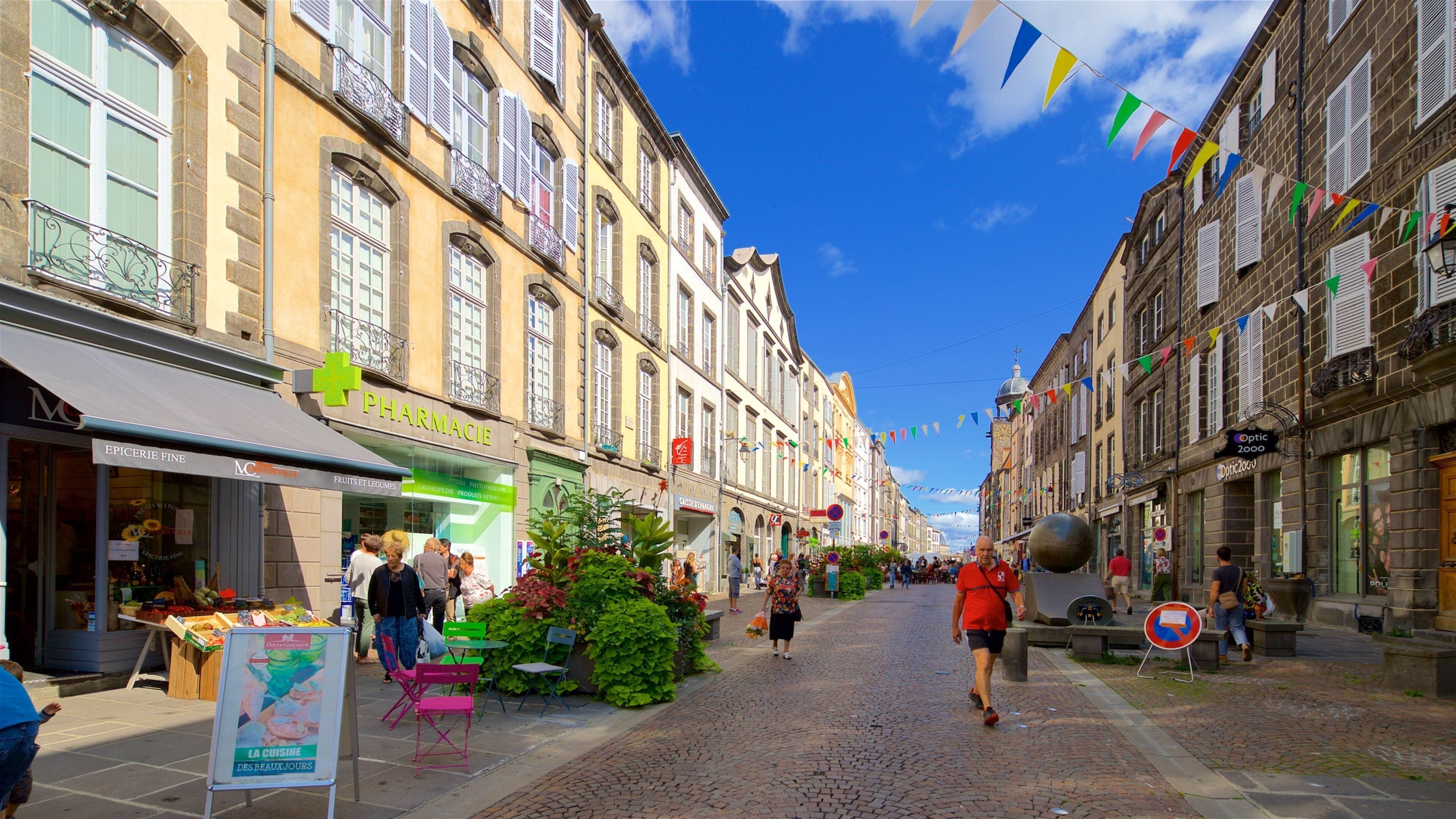 A cobblestone street lined with historic buildings, a pharmacy, and a market stall with fresh produce.